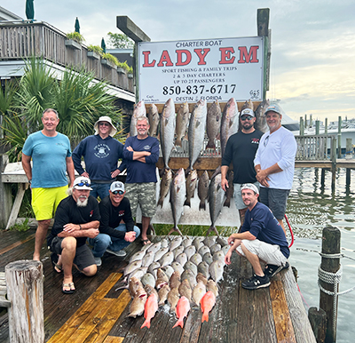 gallery-example A group of nine people pose on a dock in front of a Lady Em charter boat sign, with a display of large fish hanging and more fish laid out on the wooden dock, with water and buildings in the background.