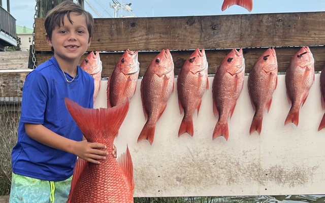 home-block-boy-with-many-fish A smiling young boy in a blue shirt holds a large red fish, standing next to a display board lined with several similar red fish, all hanging by their tails.