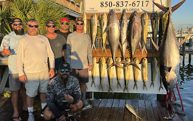 home-block-catch-pose Six men pose on a dock in front of a display of large fish they caught, hanging on hooks with a sign reading Destin, Florida and a phone number above. The sun is shining, creating warm, golden lighting.