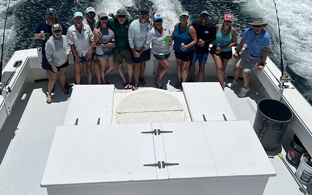 home-block-corporate-trips A group of twelve people stand close together, smiling on the deck of a boat out at sea, with fishing rods and waves visible in the background under bright daylight.
