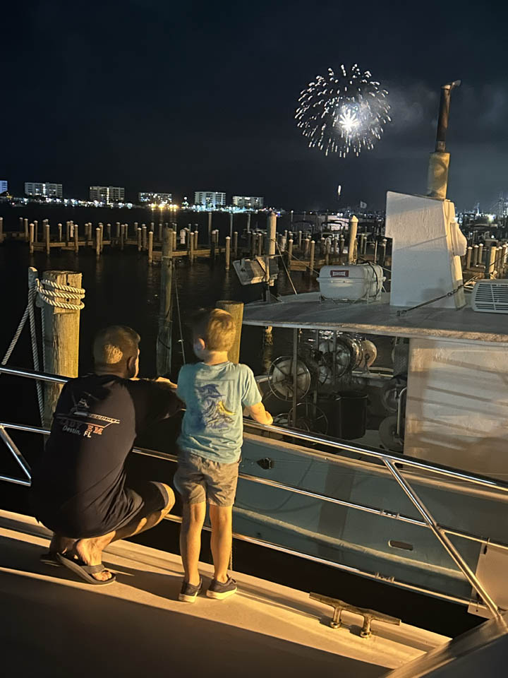 gallery-main-10 Lady EM Design Florida Charters - A man and a young boy stand on a boat at night, looking out over a marina as fireworks light up the sky in the distance. The marina is filled with other boats and city lights are visible in the background.