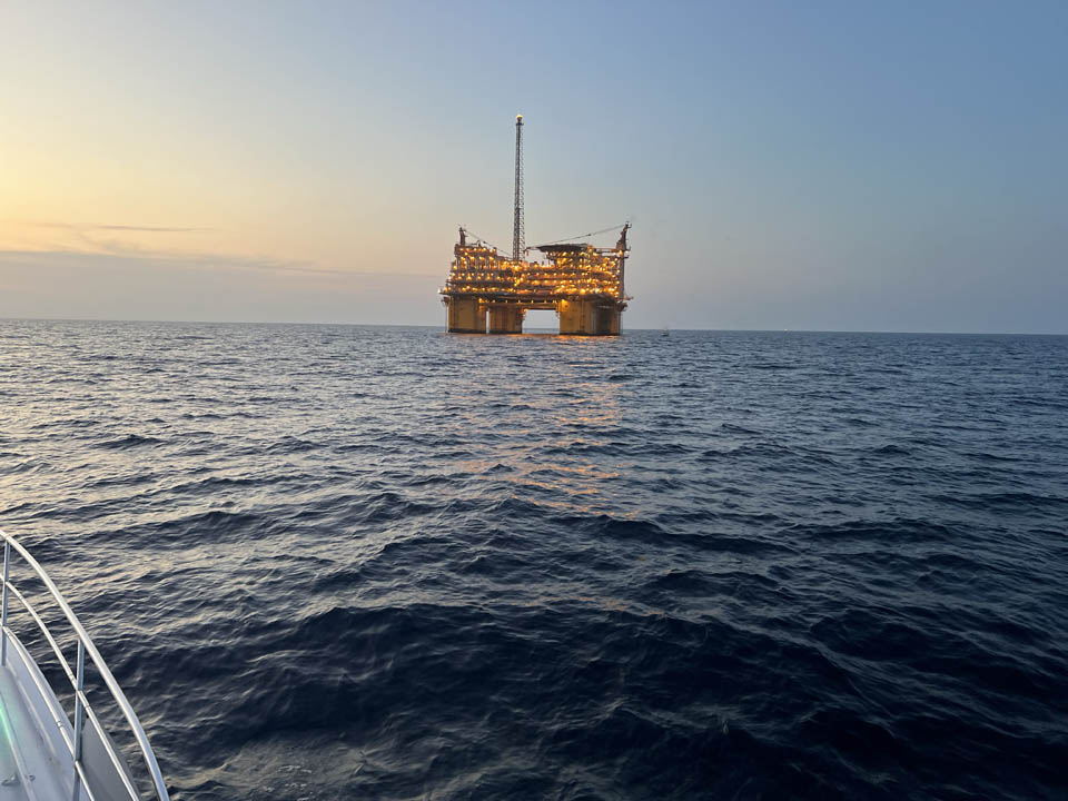 gallery-main-12 Lady EM Design Florida Charters - An offshore oil platform illuminated with lights stands in the middle of the ocean at dusk, with calm waves and a boat’s edge visible in the foreground. The sky is clear with a slight gradient from sunset.