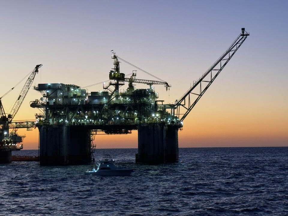 gallery-main-14 Lady EM Design Florida Charters - An offshore oil rig illuminated with lights stands in the ocean at sunset, with cranes visible and a small boat nearby on the water.