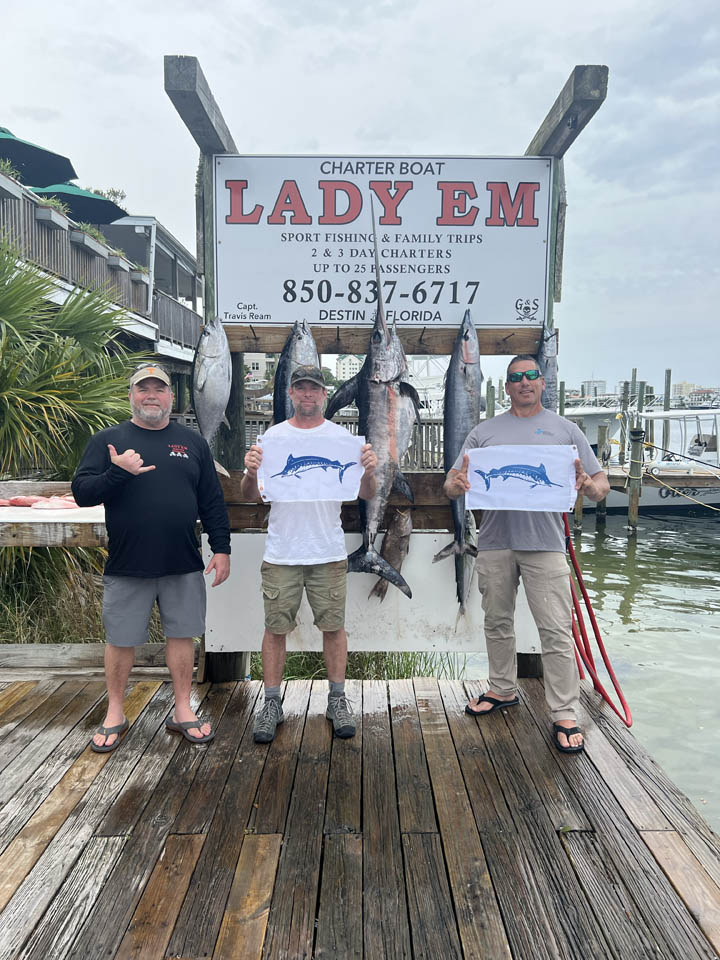 gallery-main-26 Three men stand on a dock in front of a Lady Em charter boat sign, displaying large fish they caught. Two men hold t-shirts with a marlin graphic. The marina and boats are visible in the background.
