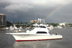 ladyem-glamour-shot-3 A white fishing boat with a red stripe floats on the water near a marina, with buildings and other boats in the background. A faint rainbow appears in the cloudy sky above.